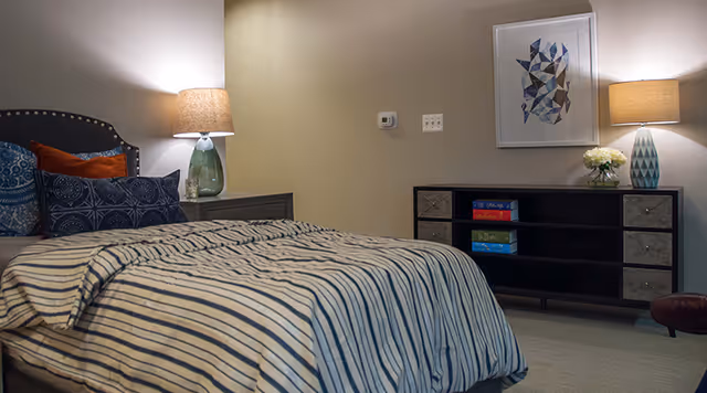 A bedroom with a bed covered in a striped comforter and multiple pillows. There are two bedside tables with lamps on either side of the bed. Across from the bed is a black dresser with drawers and shelves, holding board games and a vase with flowers. A framed abstract artwork hangs on the wall above the dresser.