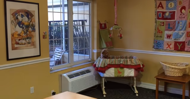 A cozy room with yellow walls featuring a framed picture of Donald Duck on the left wall and a colorful alphabet quilt hanging on the right wall. Below the quilt is a wooden table with a wicker basket. In the corner near a window is a baby bassinet with a teddy bear inside. Outside the window, part of a patio with chairs and a railing is visible.