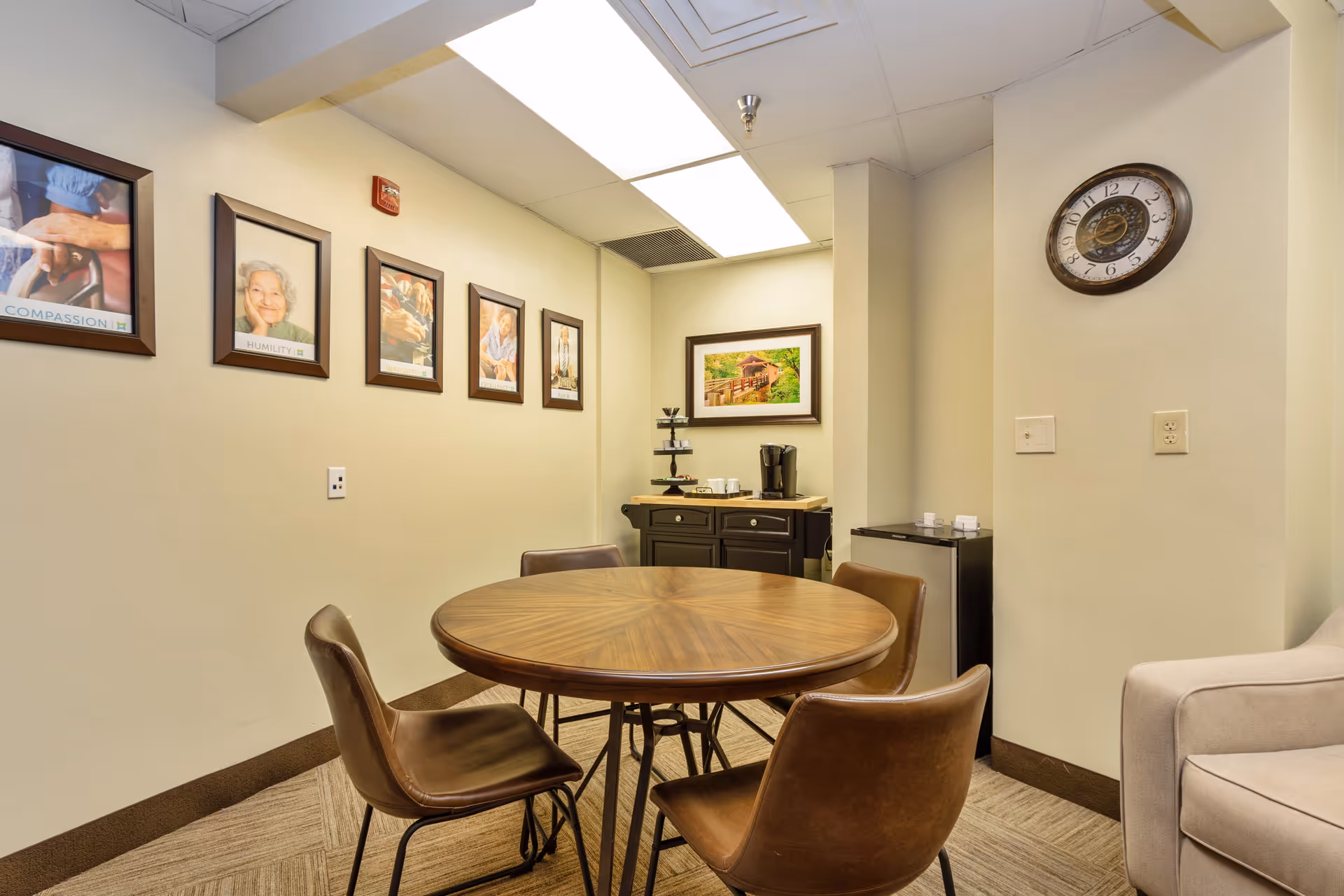 A small seating area with a round wooden table surrounded by four brown chairs. On the wall are five framed pictures, including portraits and a landscape. A black cabinet with a coffee maker and cups is in the corner, and a clock is mounted on the adjacent wall. The room has beige walls and carpeted flooring.
