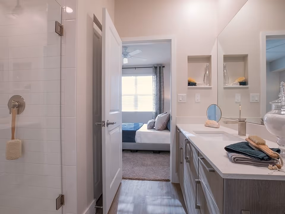 View of a modern bathroom with a glass shower door on the left, a white door leading to a bedroom with a bed and window in the background, and a long vanity with a large mirror, sink, and neatly folded towels on the right.