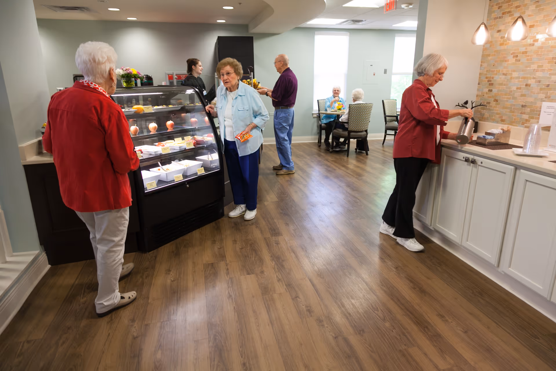 Several elderly people in a common area of a senior living facility. One woman in a red jacket stands near a display case with food items, another woman in a light blue jacket is selecting something from the case, a man in a purple shirt is standing near the back, and another woman in a red shirt is pouring a drink at a counter. In the background, two elderly people are seated at a table near windows.