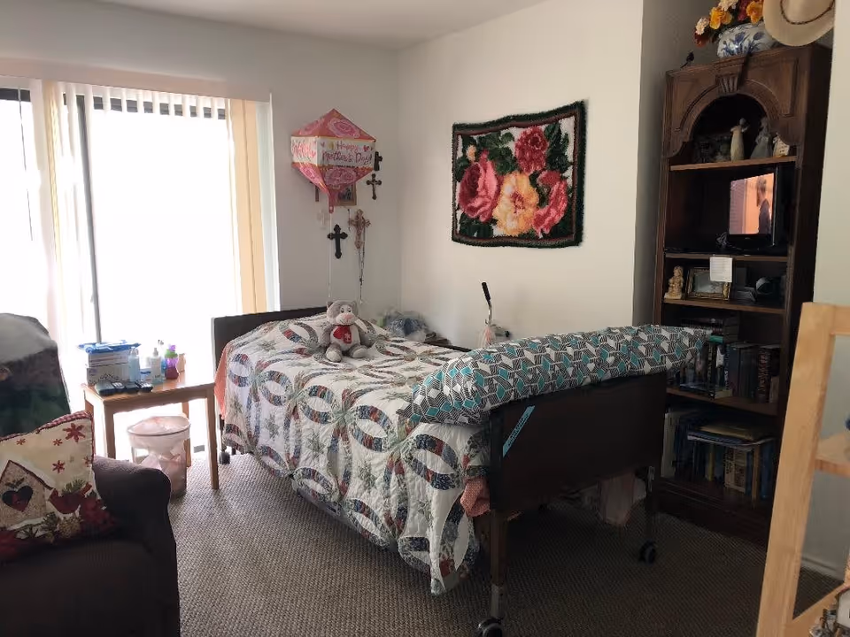 A cozy bedroom with a single bed covered in a patterned quilt and a stuffed animal on top. There is a wooden bookshelf with a small TV, books, and decorative items. A floral wall hanging and two crosses are displayed on the wall. A small wooden table next to the bed holds various personal items, and a sliding glass door with vertical blinds lets in natural light. A cushioned armchair with a decorative pillow is partially visible.