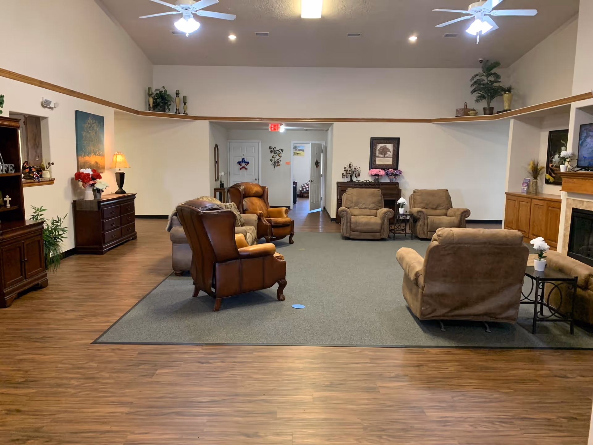 A spacious senior living facility common area with several cushioned armchairs arranged on a large gray rug. The room has wood flooring, a ceiling fan with lights, decorative plants, framed artwork on the walls, and a fireplace with a TV mounted above it. There are doorways leading to other rooms in the background.