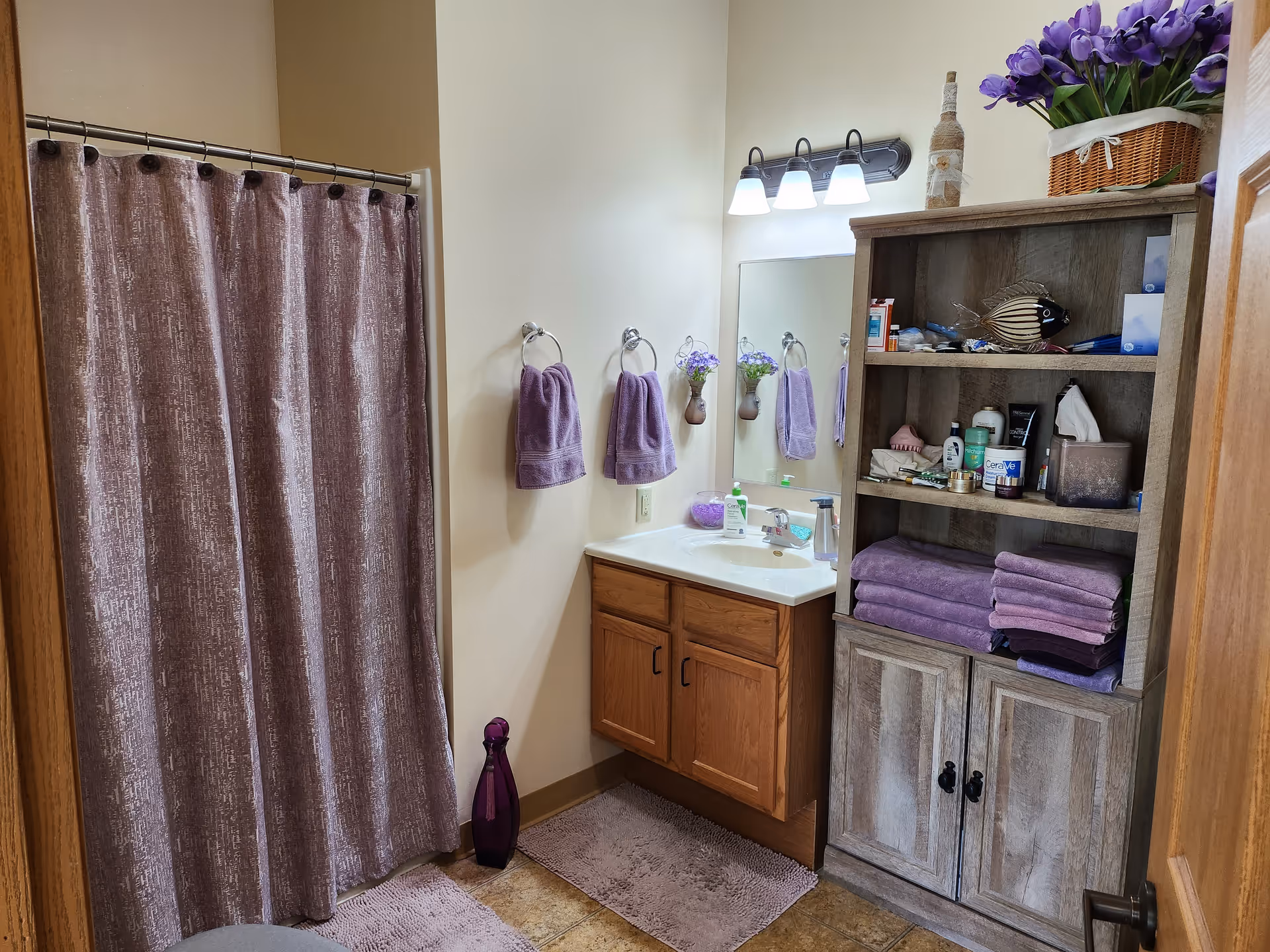 A bathroom with a shower covered by a purple curtain, a wooden vanity with a sink and mirror above it, and a wooden shelving unit filled with folded purple towels and various toiletries. The walls are light-colored, and there are purple hand towels hanging on the wall next to the sink. The floor has brown tiles and matching purple bath mats.