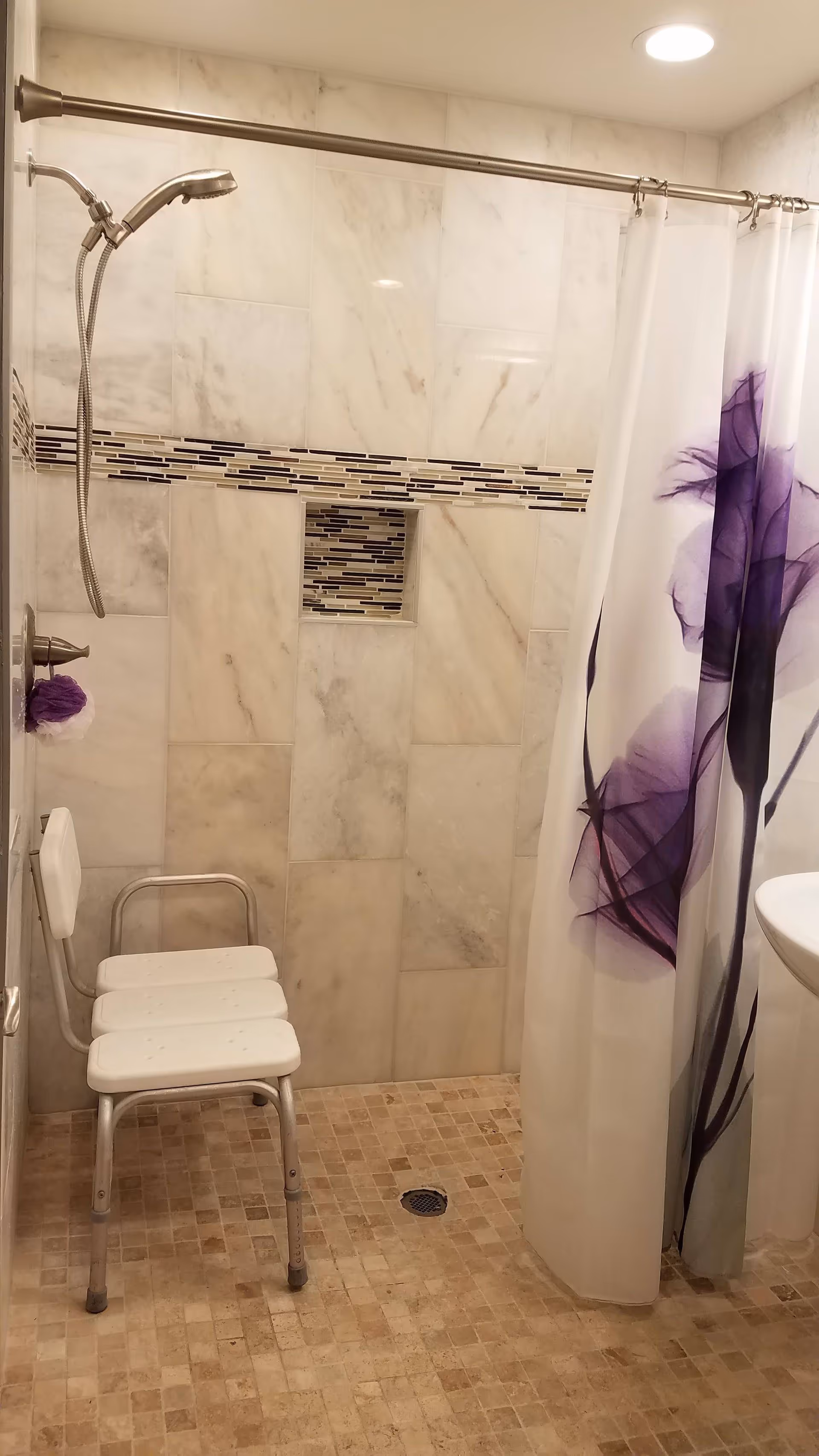 A bathroom shower area with beige tiled floor and marble-patterned wall tiles. There is a silver showerhead attached to the wall, a white shower chair with metal legs, and a white shower curtain with purple floral designs. A small recessed shelf with mosaic tiles is built into the wall.