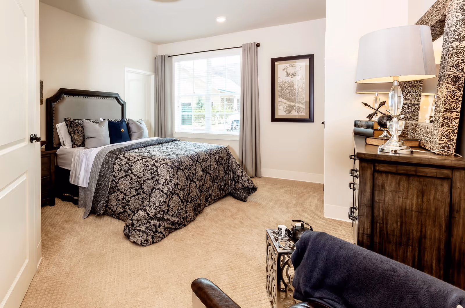 A neatly made bed with a dark patterned comforter and multiple pillows in a bright bedroom. The room features beige carpeting, a window with gray curtains, a framed picture on the wall, a wooden dresser with a decorative mirror and lamp, and a small side table with a teapot and cup. A dark upholstered chair is partially visible in the foreground.