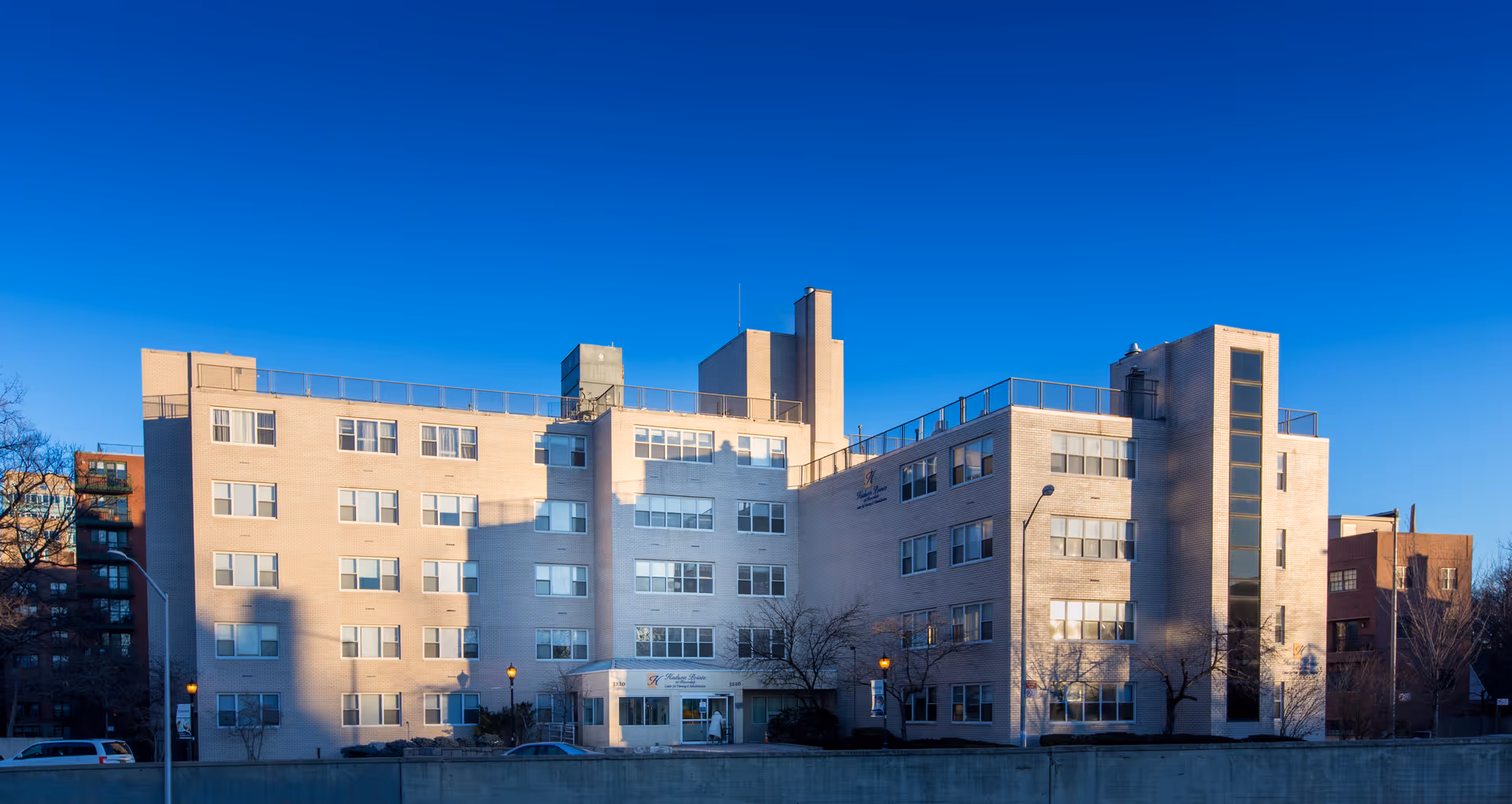 Front exterior of a multi-story beige-brick nursing and rehabilitation building against a clear blue sky.