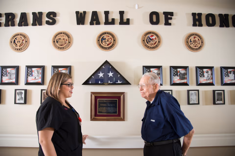 A woman and an elderly man stand and talk in front of a Veterans Wall of Honor displaying military emblems, framed photos, and a folded American flag in a triangular case.