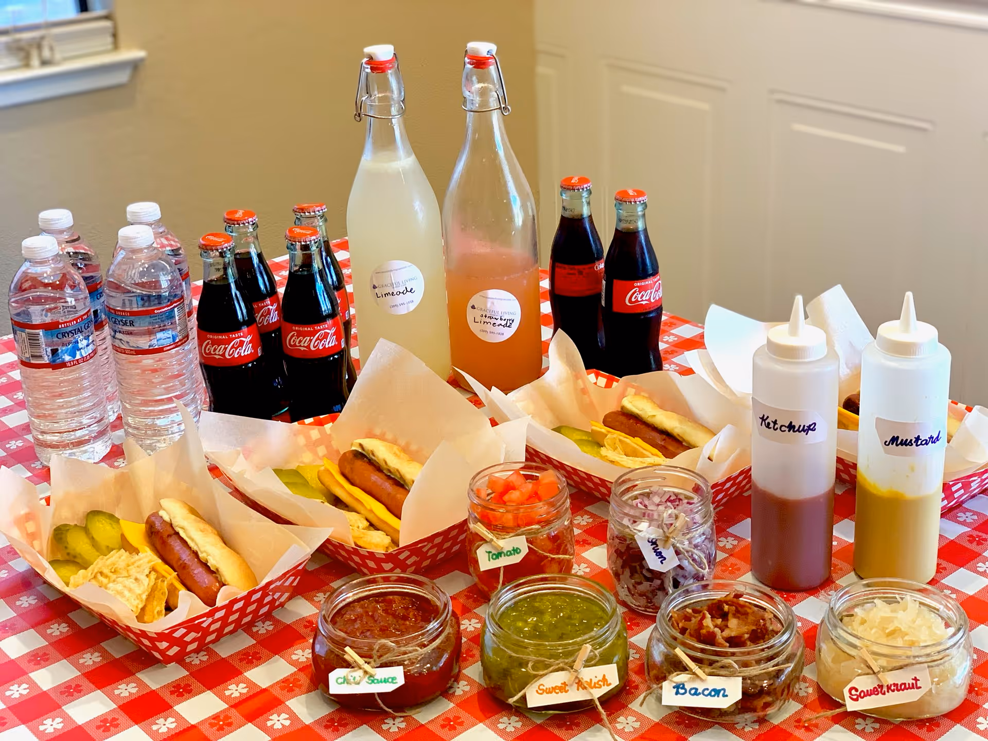 A table with a red and white checkered tablecloth displaying three trays each containing a hot dog with pickles and chips. In front of the trays are jars labeled chili sauce, sweet relish, tomato, onion, bacon, and sauerkraut. There are also squeeze bottles labeled ketchup and mustard. Behind the trays are bottles of water, glass bottles of Coca-Cola, and two large glass bottles labeled lemonade and strawberry lemonade.