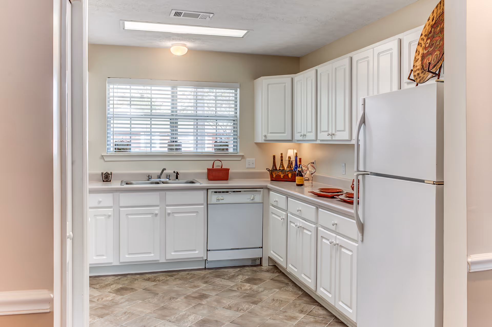 A bright kitchen with white cabinets and countertops, a white refrigerator, a dishwasher, and a window with blinds above the sink. The countertop has decorative items including bottles and plates, and the floor has a tile pattern.