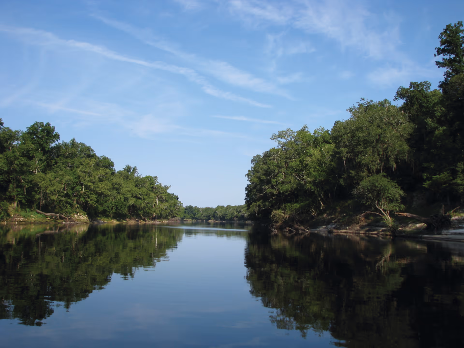 A calm river flanked by dense green trees on both sides under a blue sky with wispy clouds, reflecting clearly on the water surface.