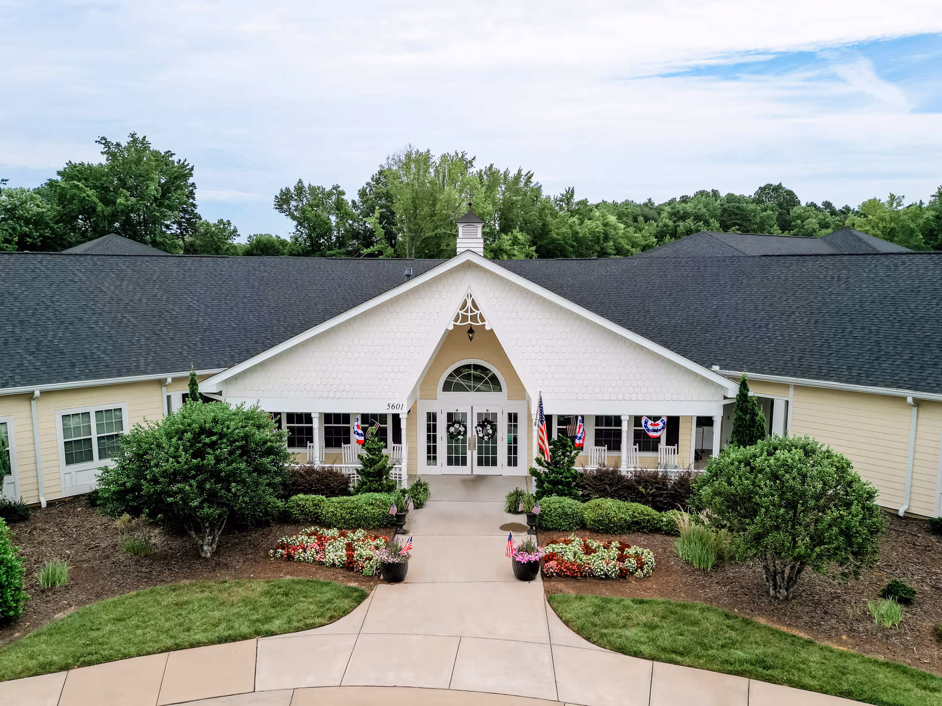 Front exterior view of a single-story senior living facility building with a peaked roof, white entrance with glass double doors, decorative wreaths, American flags, and well-maintained landscaping including bushes, flowers, and a curved walkway.