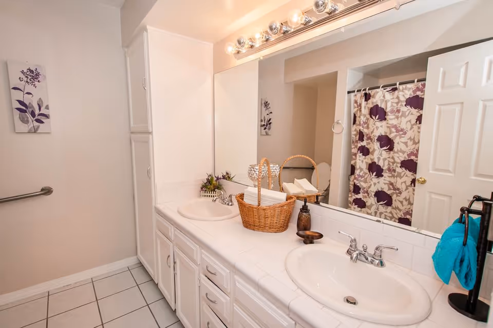 A clean bathroom with a double sink vanity featuring white countertops and cabinets. Above the sinks is a large mirror with a row of lights. There is a wicker basket with folded towels on the counter, a soap dispenser, and a small decorative plant. A shower curtain with a purple floral pattern is visible in the background, along with a white door and a towel rack holding a blue towel.