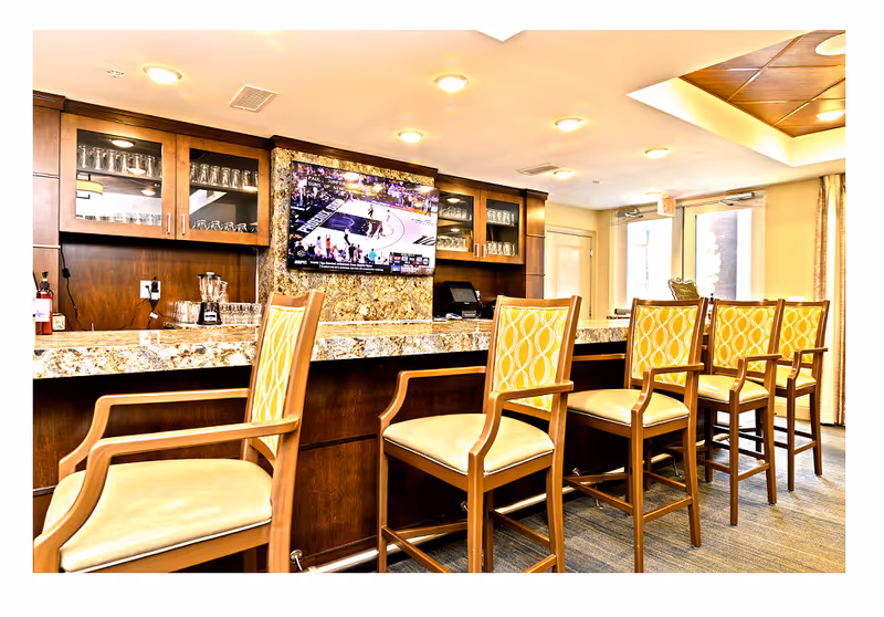 Interior view of a bar area with a granite countertop and five wooden bar stools with yellow patterned cushions. Behind the counter are wooden cabinets with glassware and a mounted flat-screen TV showing a basketball game. The room is well-lit with ceiling lights and has a door and windows with curtains in the background.