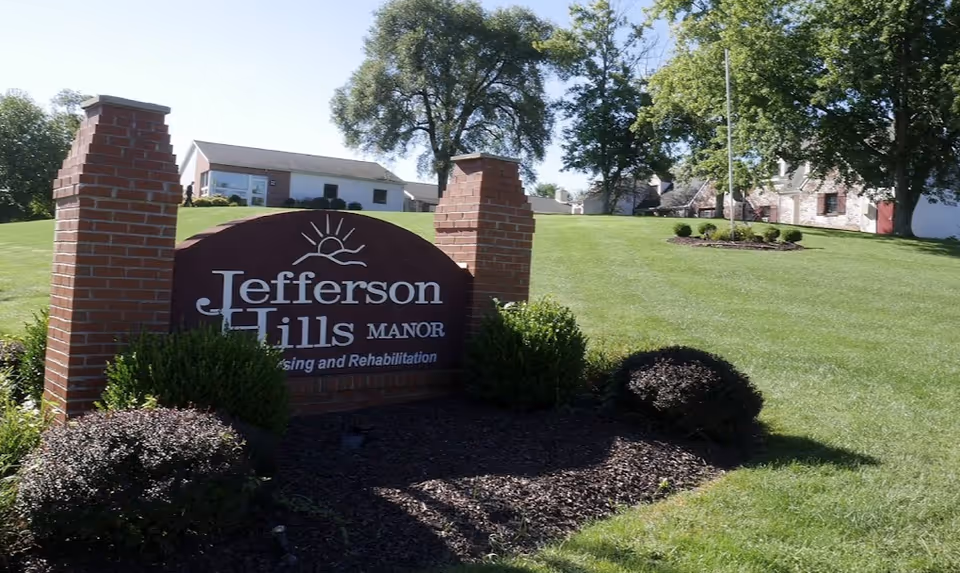 Entrance sign reading "Jefferson Hills Manor" on a landscaped lawn with buildings and trees in the background.