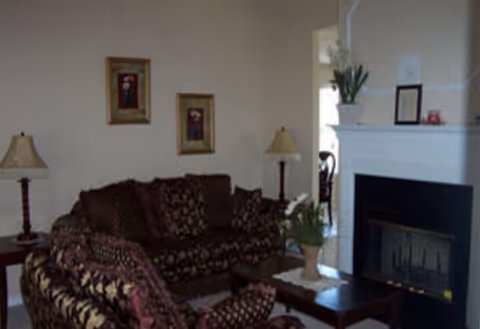 A cozy living room with two patterned sofas arranged around a dark wooden coffee table. There are two table lamps on either side of the sofas, and a white fireplace with decorative items on the mantel. Two framed floral pictures hang on the wall above one sofa.