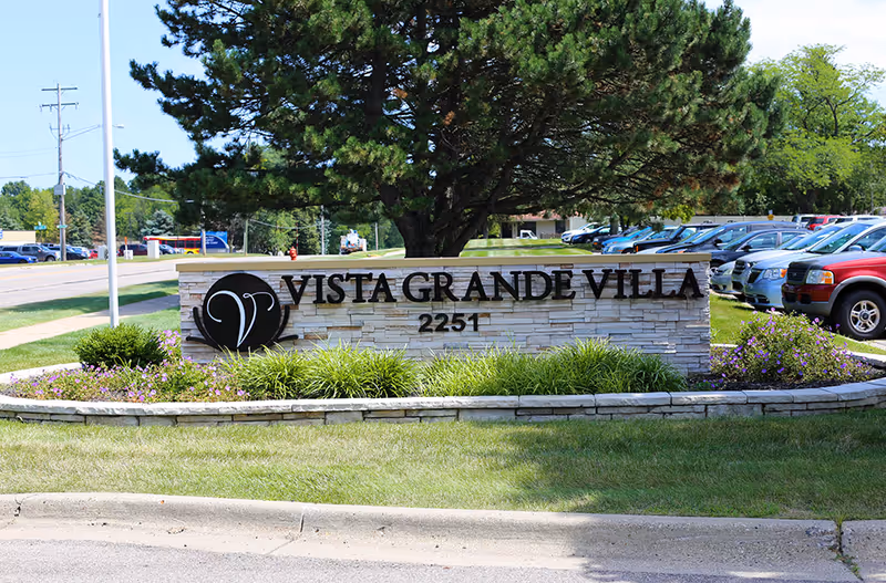 Vista Grande Villa entrance sign with stone base, greenery, and cars in parking lot.