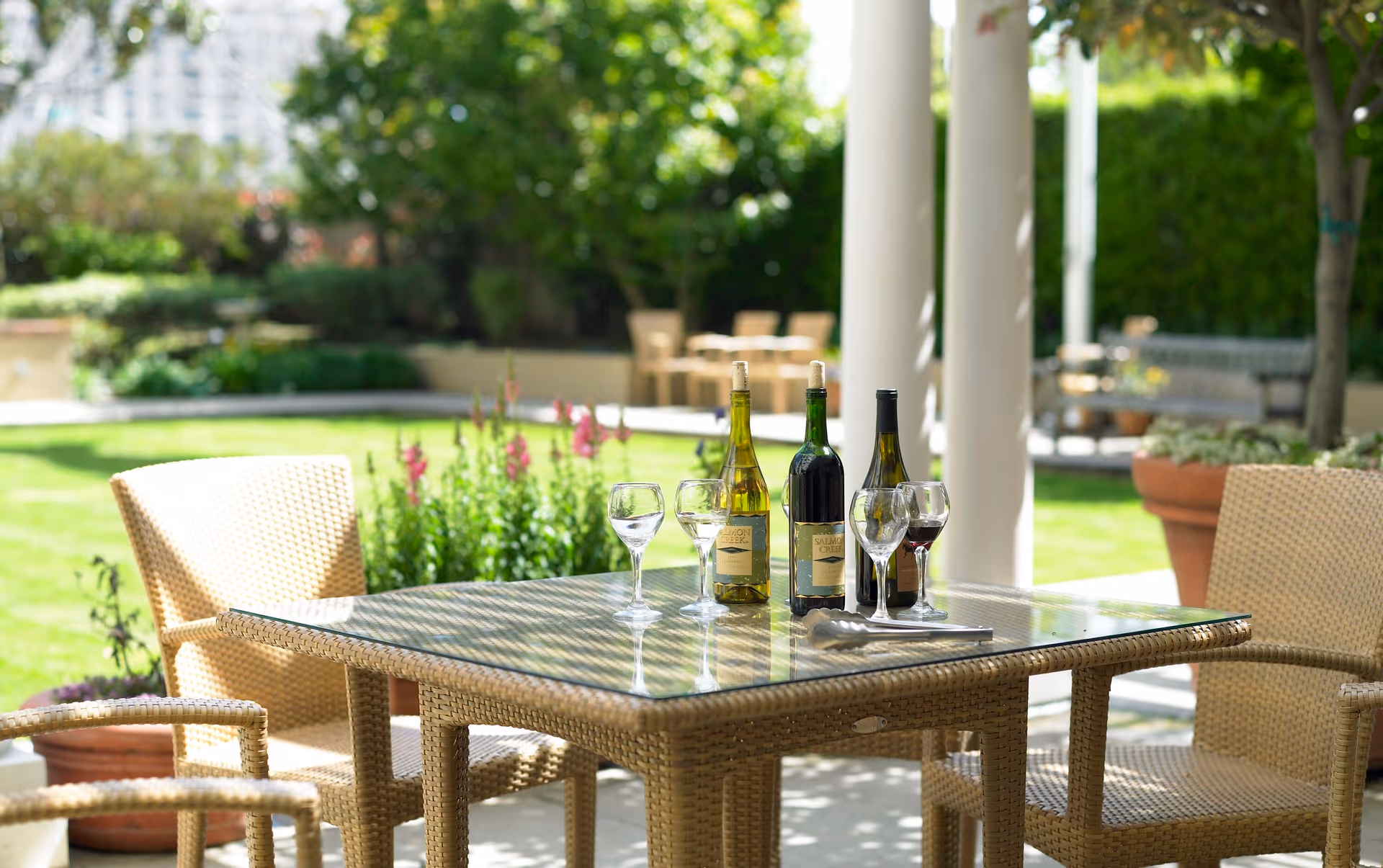 Outdoor patio with a glass-topped wicker table set with wine bottles and glasses and wicker chairs in a sunny garden.