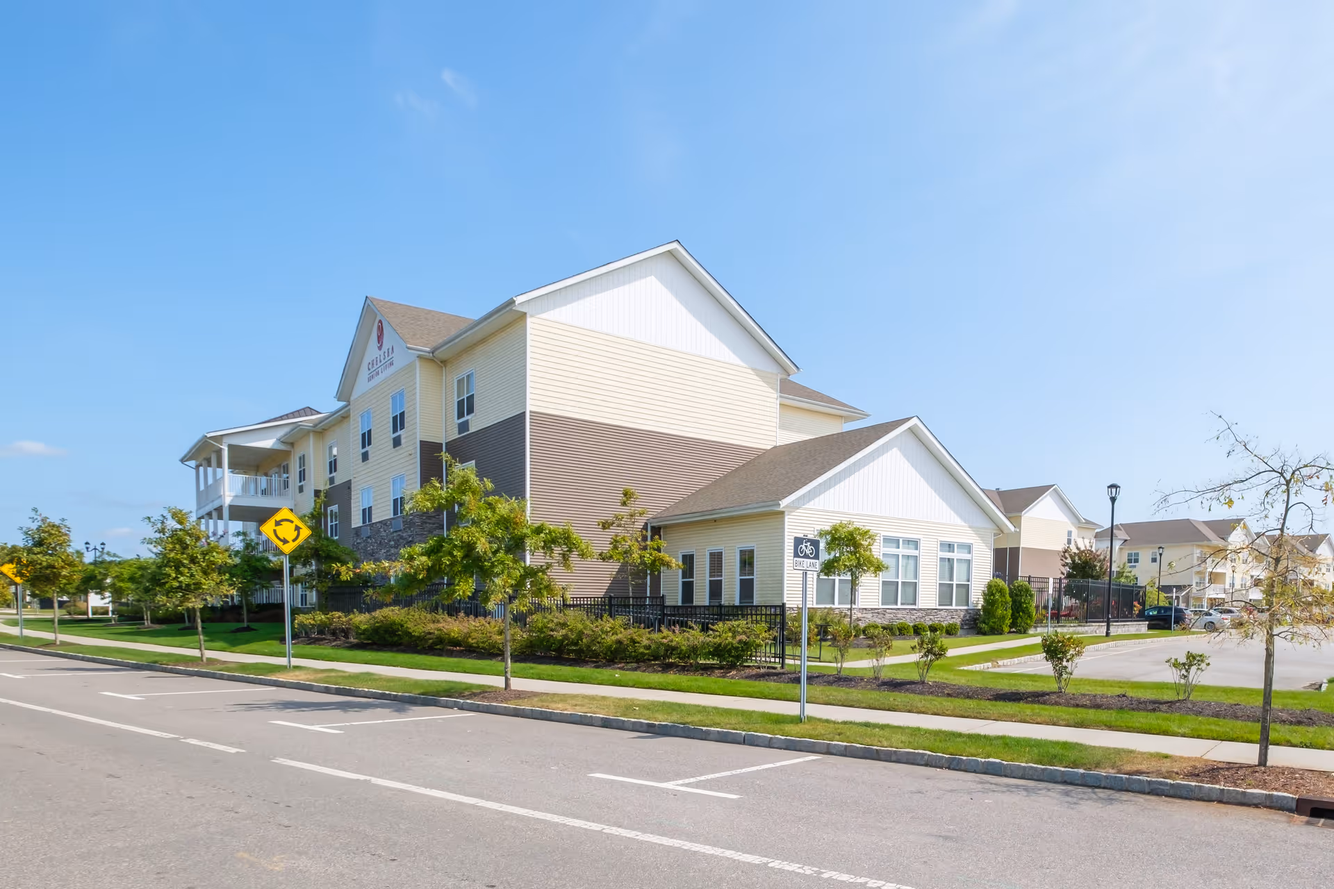 Exterior front view of The Chelsea at Brookhaven multi-story senior living building with landscaping and a clear blue sky.