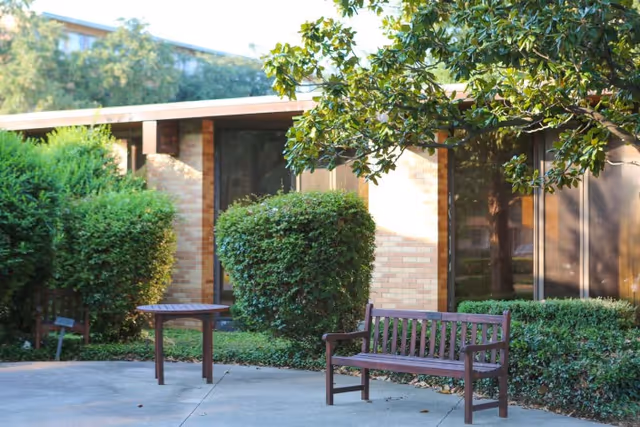 Outdoor seating area with a wooden bench and a small table on a concrete patio surrounded by green bushes and trees in front of a brick building with large windows.