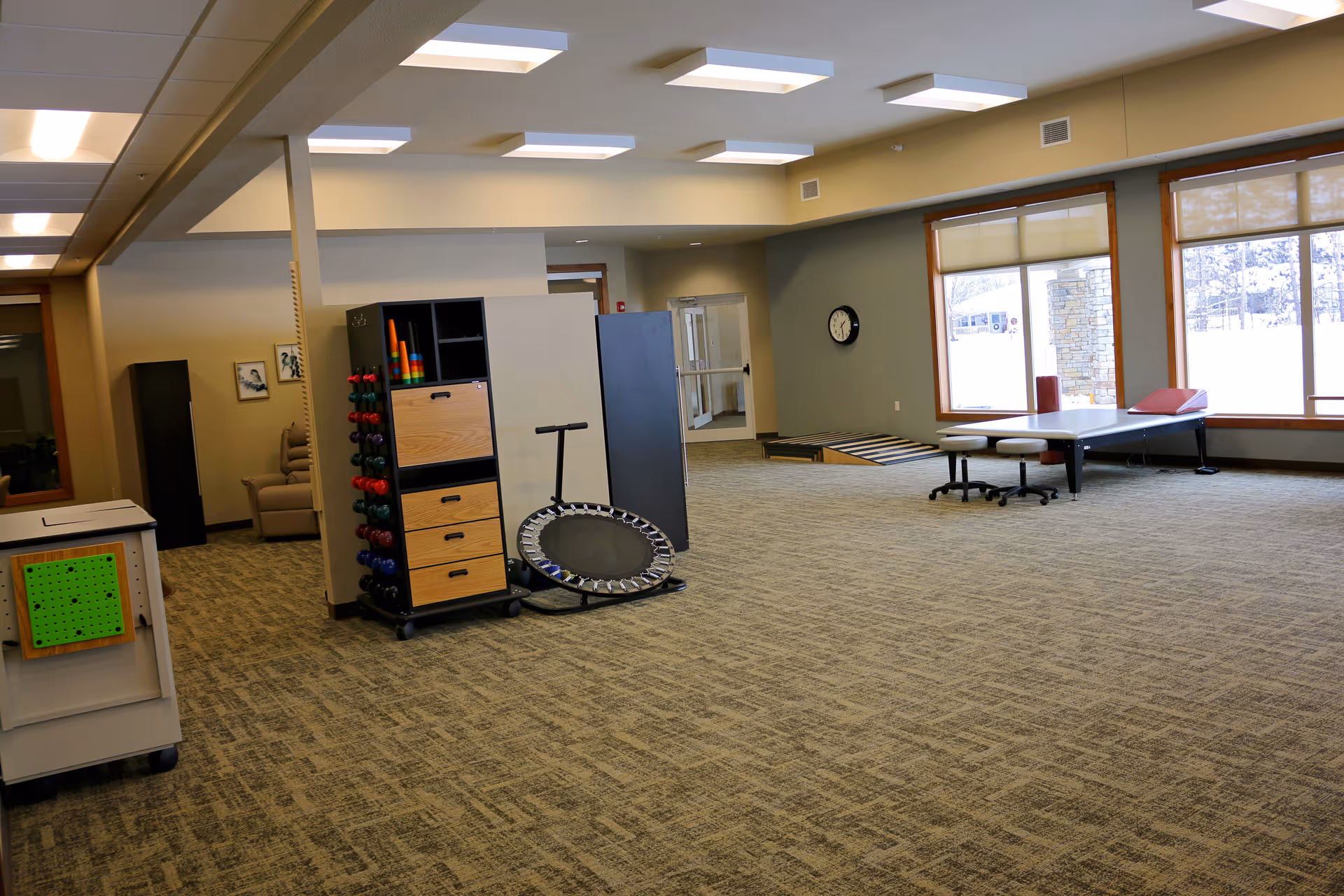 Large bright therapy/activity room with exercise equipment, a small trampoline, dumbbells, treatment table and large windows.