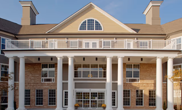 Front exterior view of a senior living facility building with large white columns, brick and beige siding, multiple windows, and a balcony above the entrance under a clear blue sky.