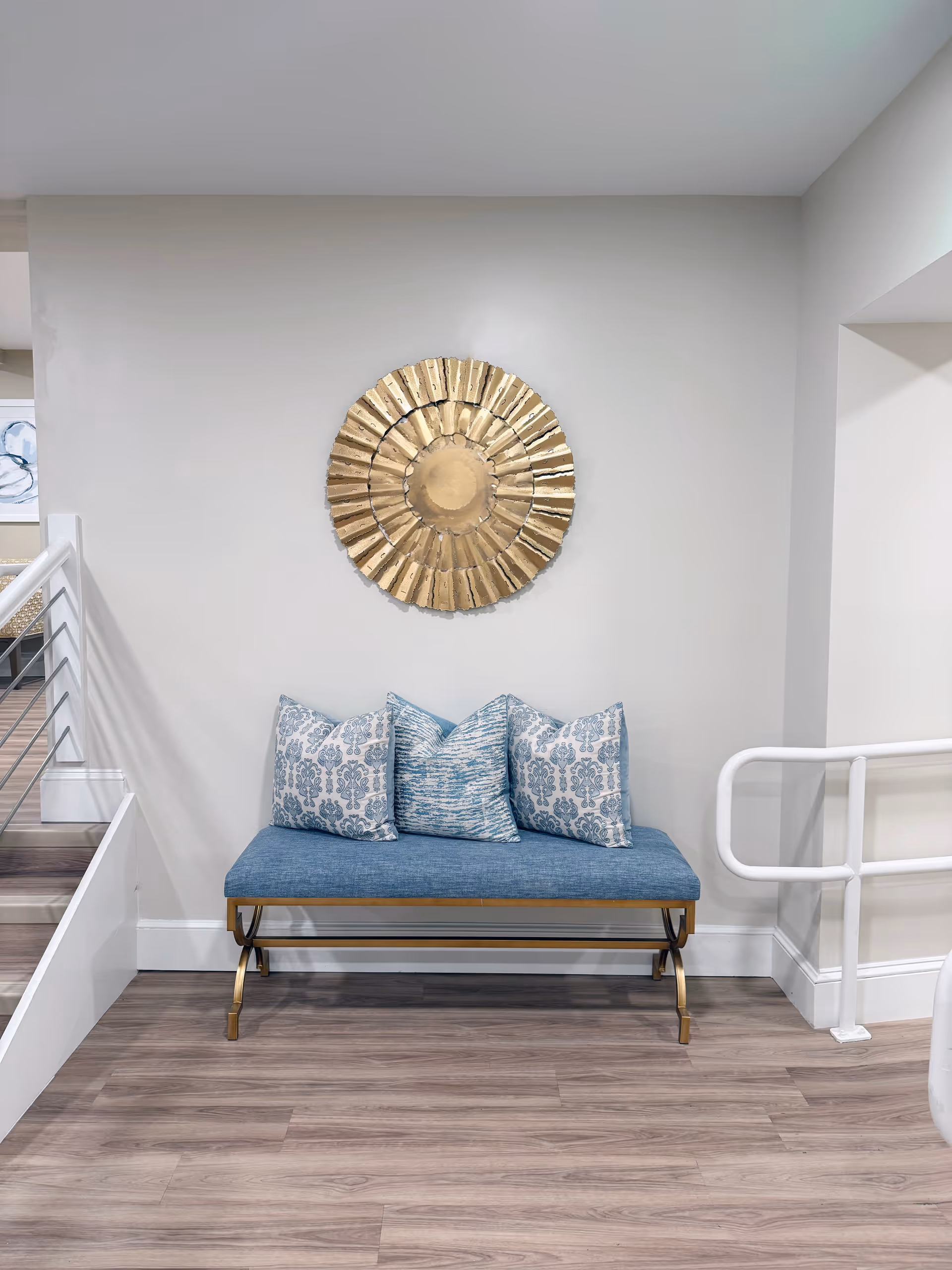 A blue cushioned bench with three decorative pillows in blue and white patterns is placed against a light gray wall. Above the bench hangs a large, round, gold decorative wall piece. The floor is wood with a light finish, and there is a white railing on the right side and stairs with a metal railing on the left.
