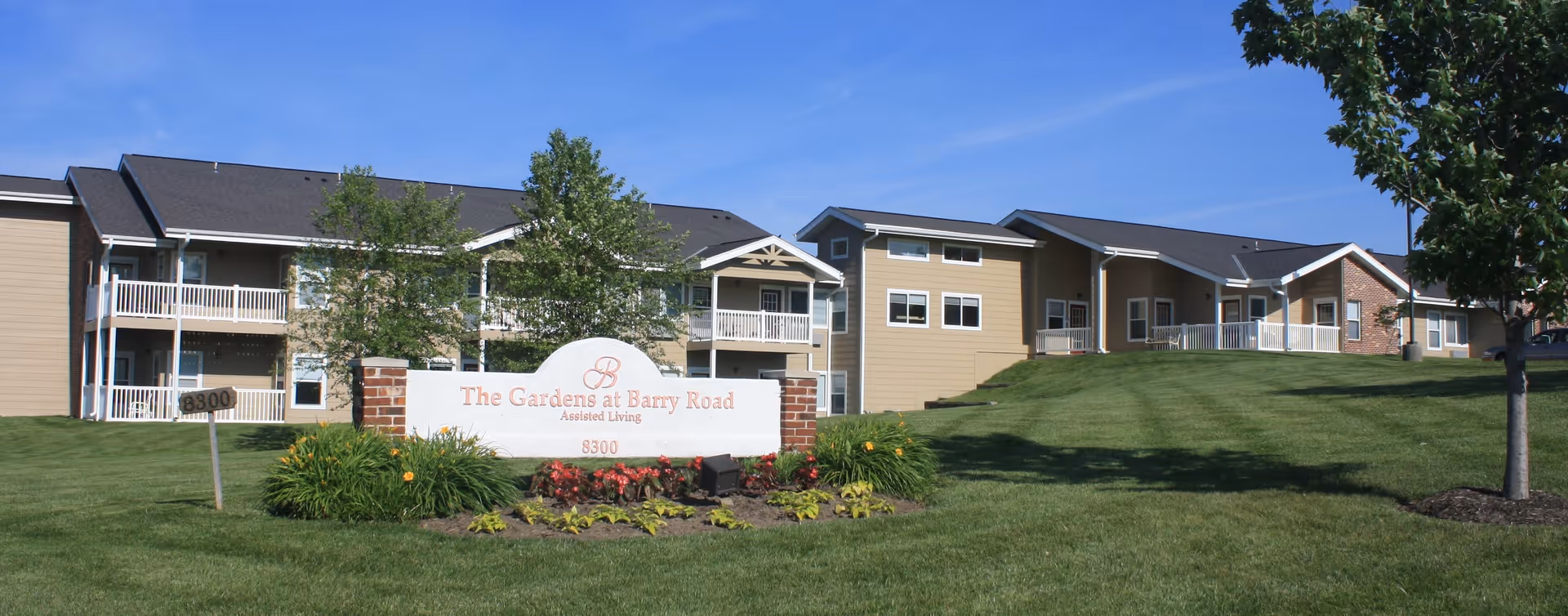 Exterior view of The Gardens at Barry Road Assisted Living and Memory Care facility showing a large, well-maintained lawn with trees and shrubs, a prominent sign with the facility name and address, and a multi-unit building with balconies under a clear blue sky.