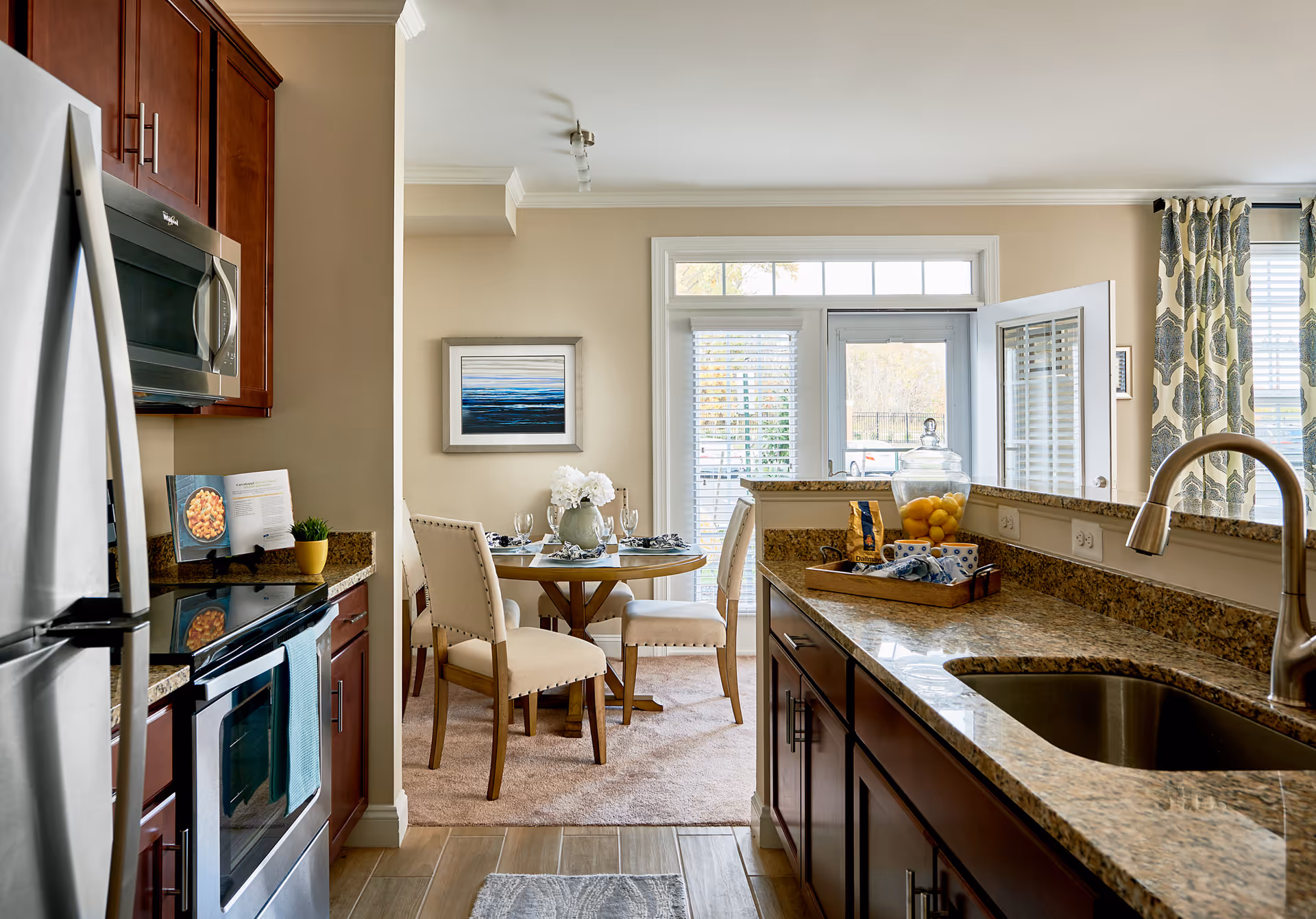 A bright kitchen and dining area in a senior living facility. The kitchen features stainless steel appliances, dark wood cabinets, and granite countertops with a sink and faucet. The dining area has a round wooden table set with four upholstered chairs, a vase with white flowers, and a framed abstract painting on the wall. Large windows and a glass door allow natural light to fill the space.