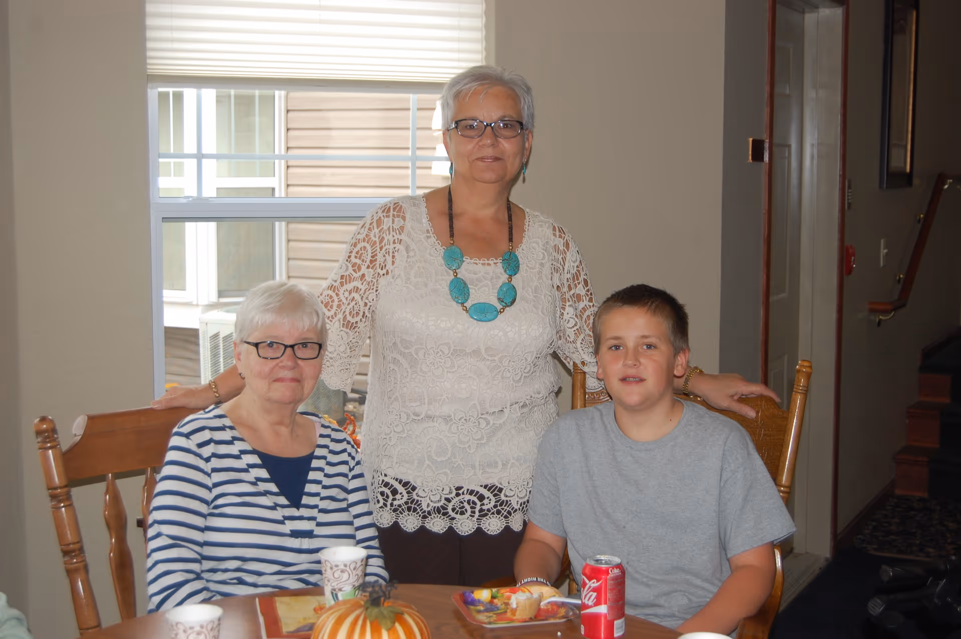 Three people gathered around a wooden dining table inside a room. An elderly woman with white hair and glasses is seated on the left wearing a striped shirt. A middle-aged woman with short gray hair, glasses, and a white lace top stands behind the table with her hands resting on the backs of two chairs. A young boy in a gray t-shirt is seated on the right. On the table are a small pumpkin decoration, paper cups, a plate with food, and a can of Coca-Cola. A window with blinds is visible in the background.
