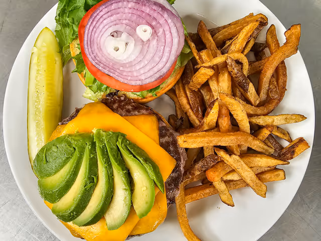 A plate with a cheeseburger topped with sliced avocado, cheddar cheese, lettuce, tomato, and a slice of red onion, served with a side of French fries and a pickle spear.
