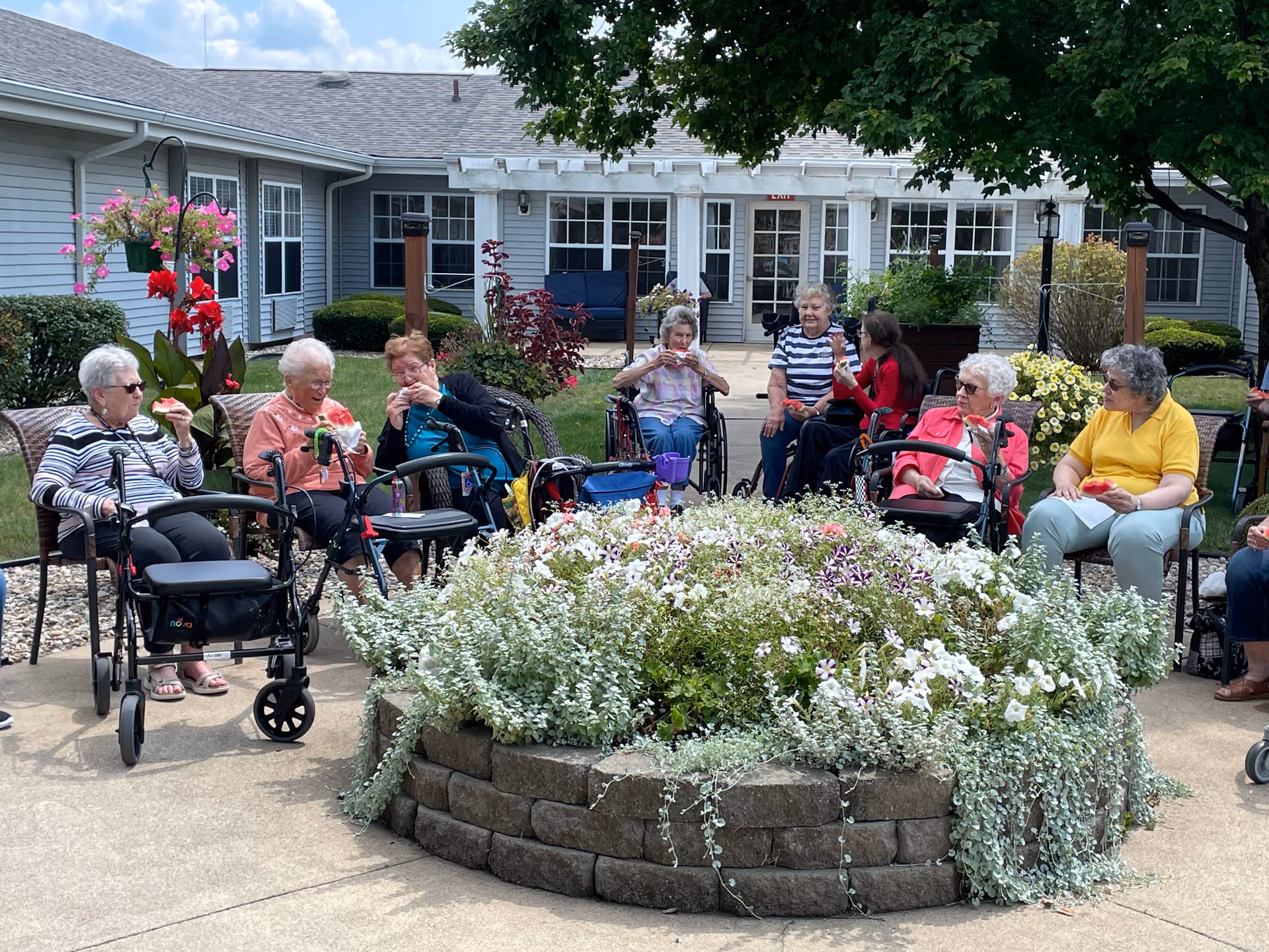 A group of elderly women sitting outdoors around a circular raised flower bed filled with white and purple flowers. Some women are seated in wheelchairs or using walkers, and they appear to be enjoying a snack together. The setting is a courtyard of a senior living facility with a building and greenery in the background under a partly cloudy sky.