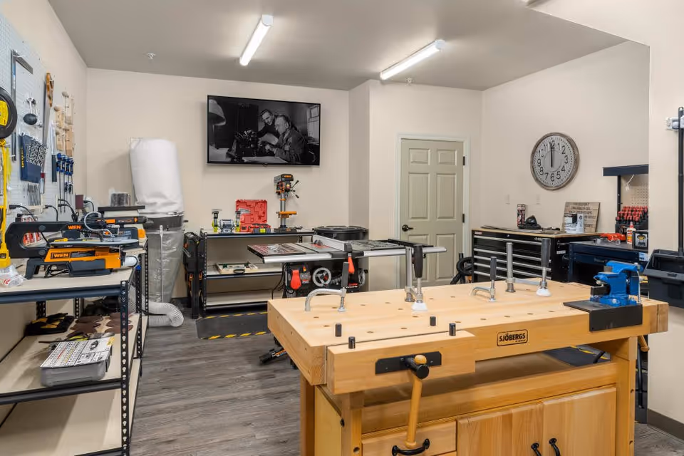 A well-organized workshop room featuring a large wooden workbench with clamps, various power tools including a drill press and saws, tool storage cabinets, and a wall-mounted black and white photograph. The room has light-colored walls and wood flooring.