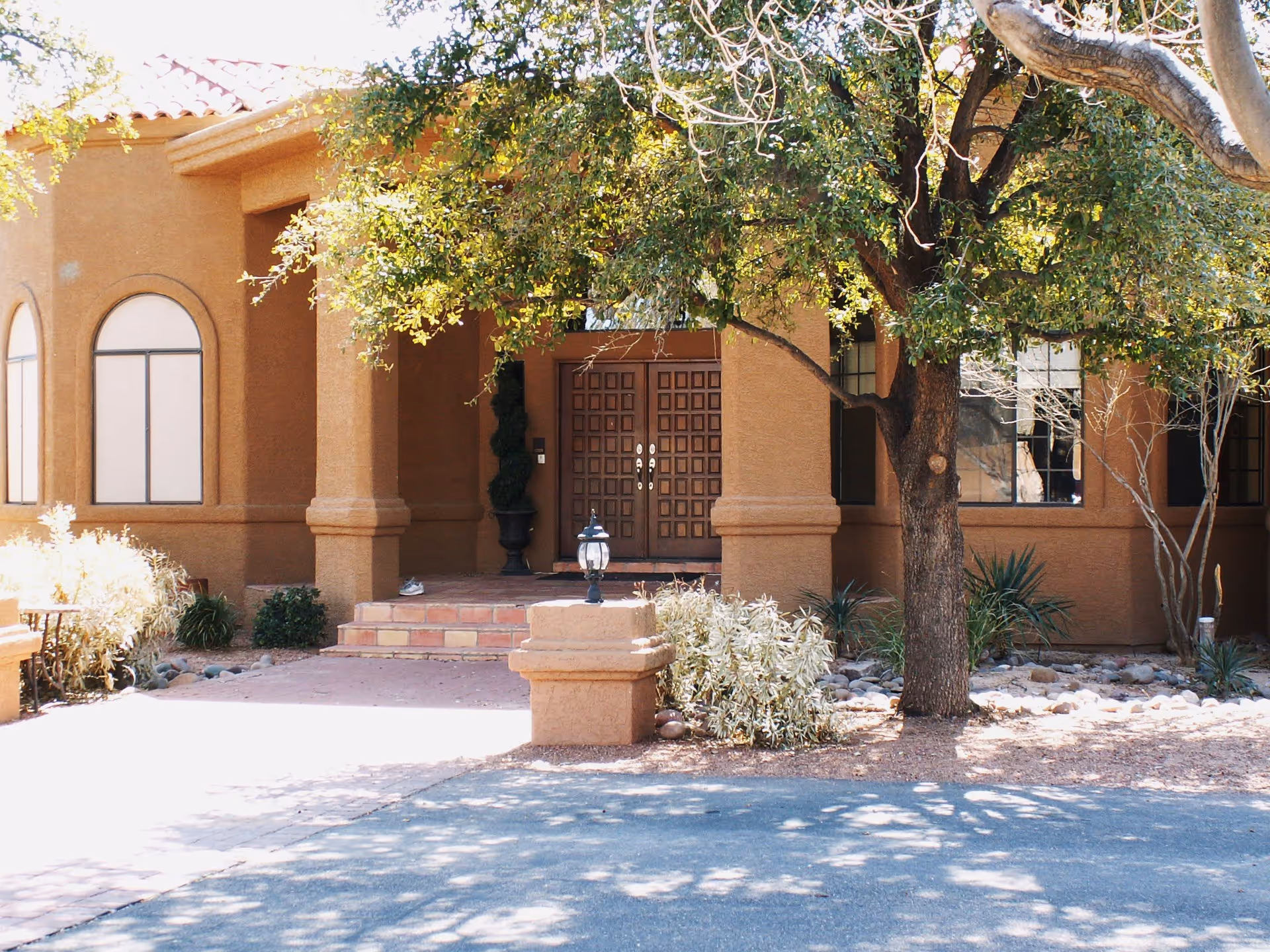 Front exterior view of a building with a brown stucco finish, arched windows, and a double wooden door entrance. There is a tree with green leaves providing shade in front of the entrance, along with some shrubs and landscaping rocks around the base of the tree.
