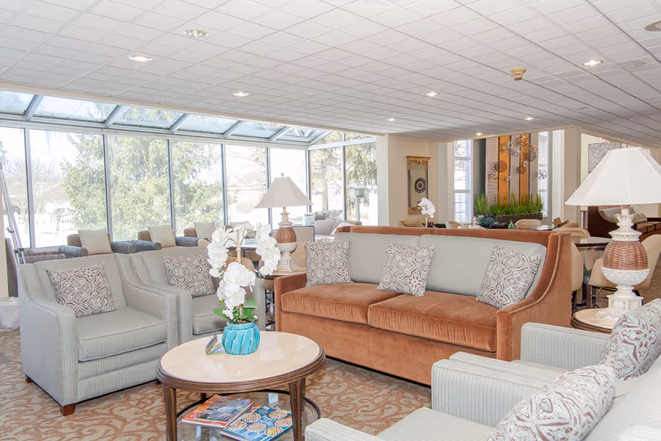 A bright and spacious living room area in a retirement community with large windows letting in natural light. The room features a brown sofa with patterned cushions, two light gray armchairs with matching cushions, a round wooden coffee table with a blue vase holding white flowers, and two table lamps with white shades. The background shows more seating and decorative elements including plants and wall art.