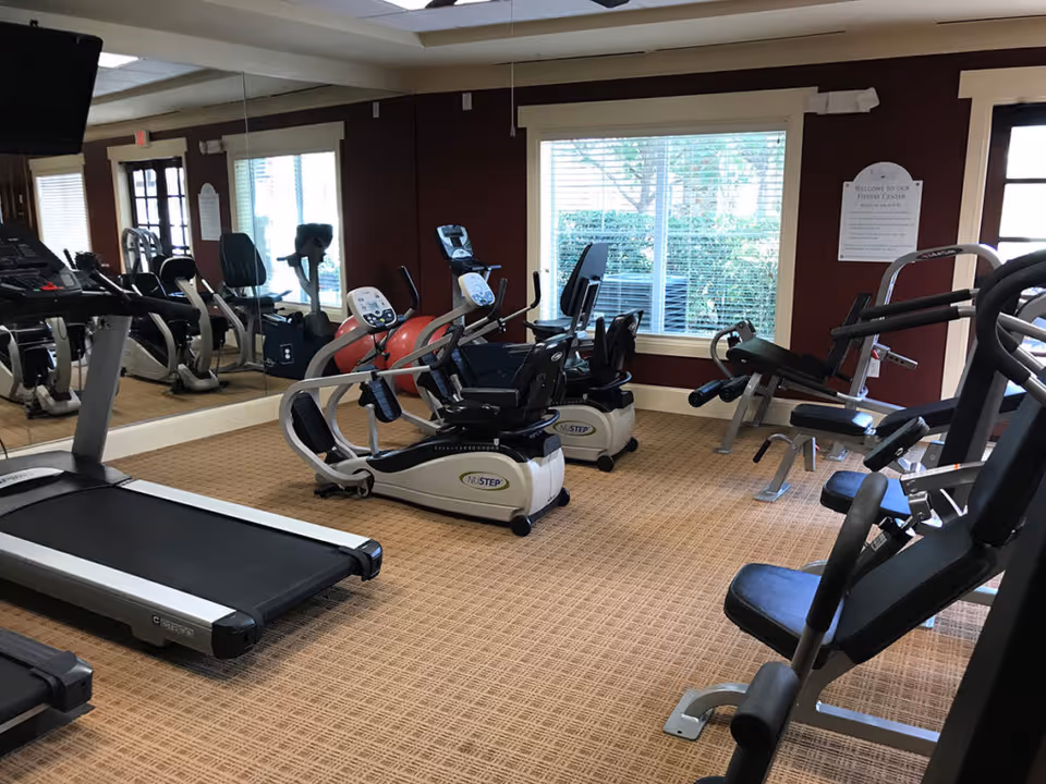 Interior view of a fitness center with exercise equipment including treadmills, stationary bikes, and weight machines. The room has carpeted flooring, large windows with blinds, and a wall mirror reflecting the equipment.