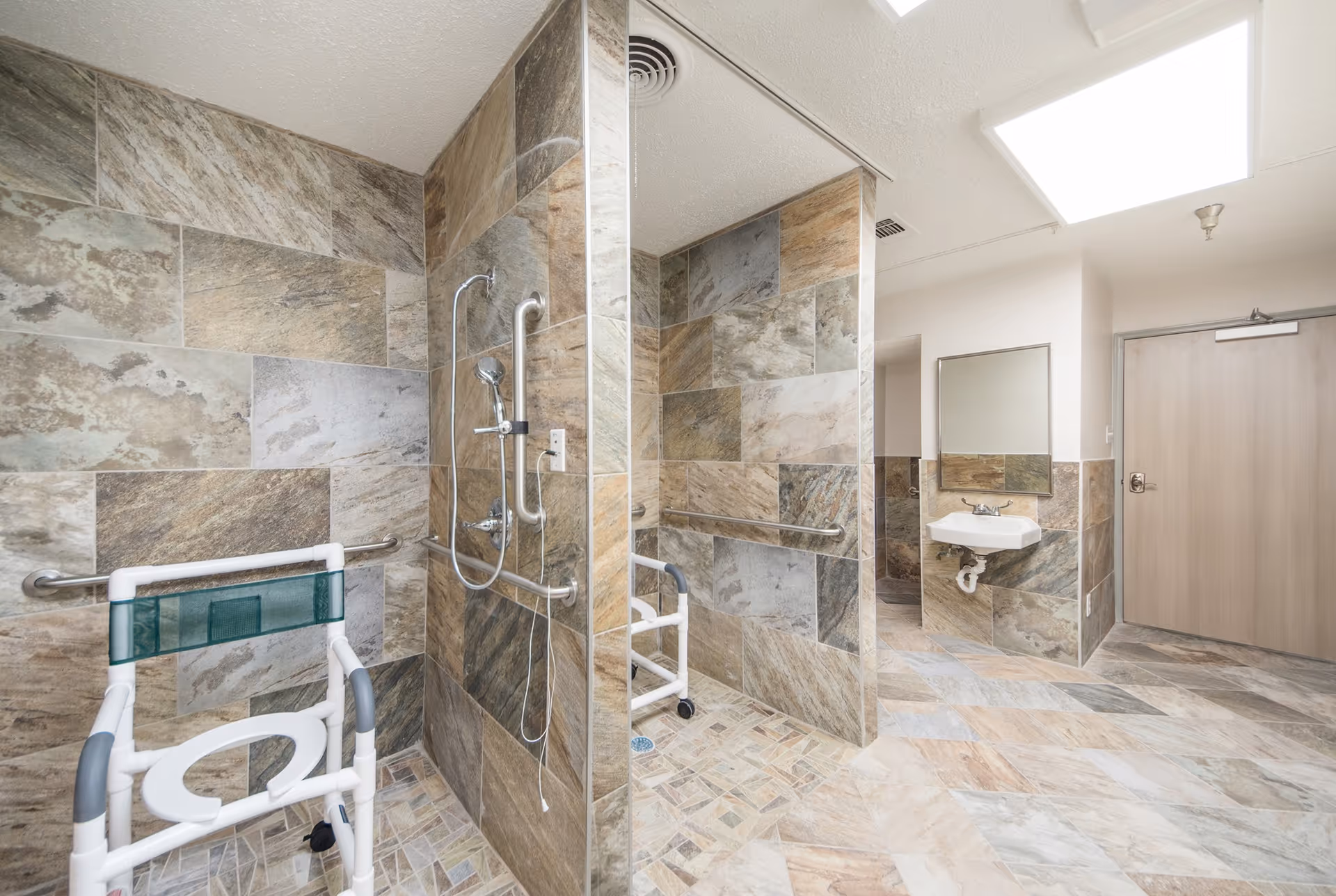 A spacious, accessible bathroom with tiled walls and floors in neutral tones. The room features a walk-in shower with grab bars and a handheld showerhead, as well as a white sink with a mirror above it. There is a shower chair with wheels positioned near the shower area. The ceiling has a skylight providing natural light, and a wooden door is visible in the background.