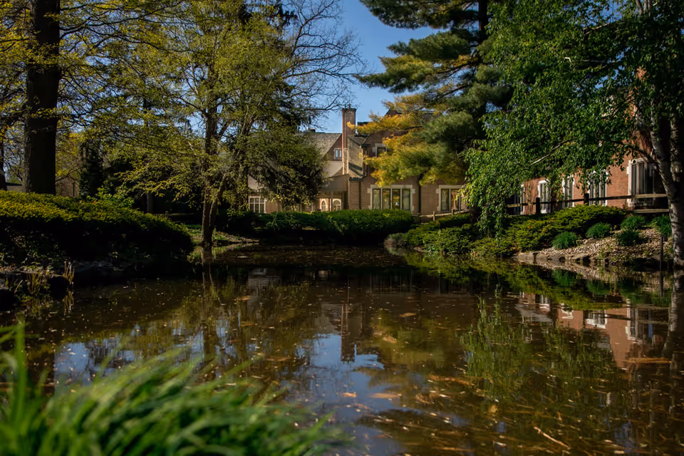A serene pond surrounded by lush green trees and bushes with a brick building partially visible in the background under a clear blue sky.
