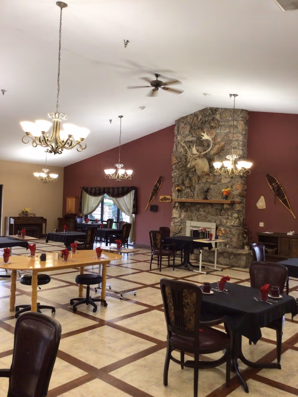 Spacious dining room with tables and chairs, chandeliers, and a large stone fireplace topped by a mounted deer head.