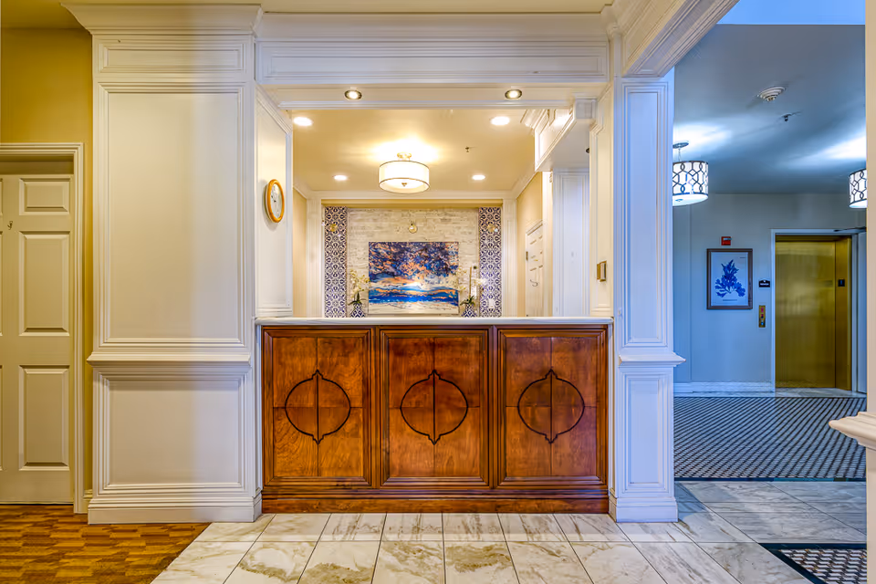 Reception area in a senior living facility featuring a wooden front desk with decorative paneling, a wall clock, and a colorful painting with floral arrangements behind the desk. To the right, there is a hallway with patterned carpet leading to a gold-colored elevator door and framed artwork on the wall.