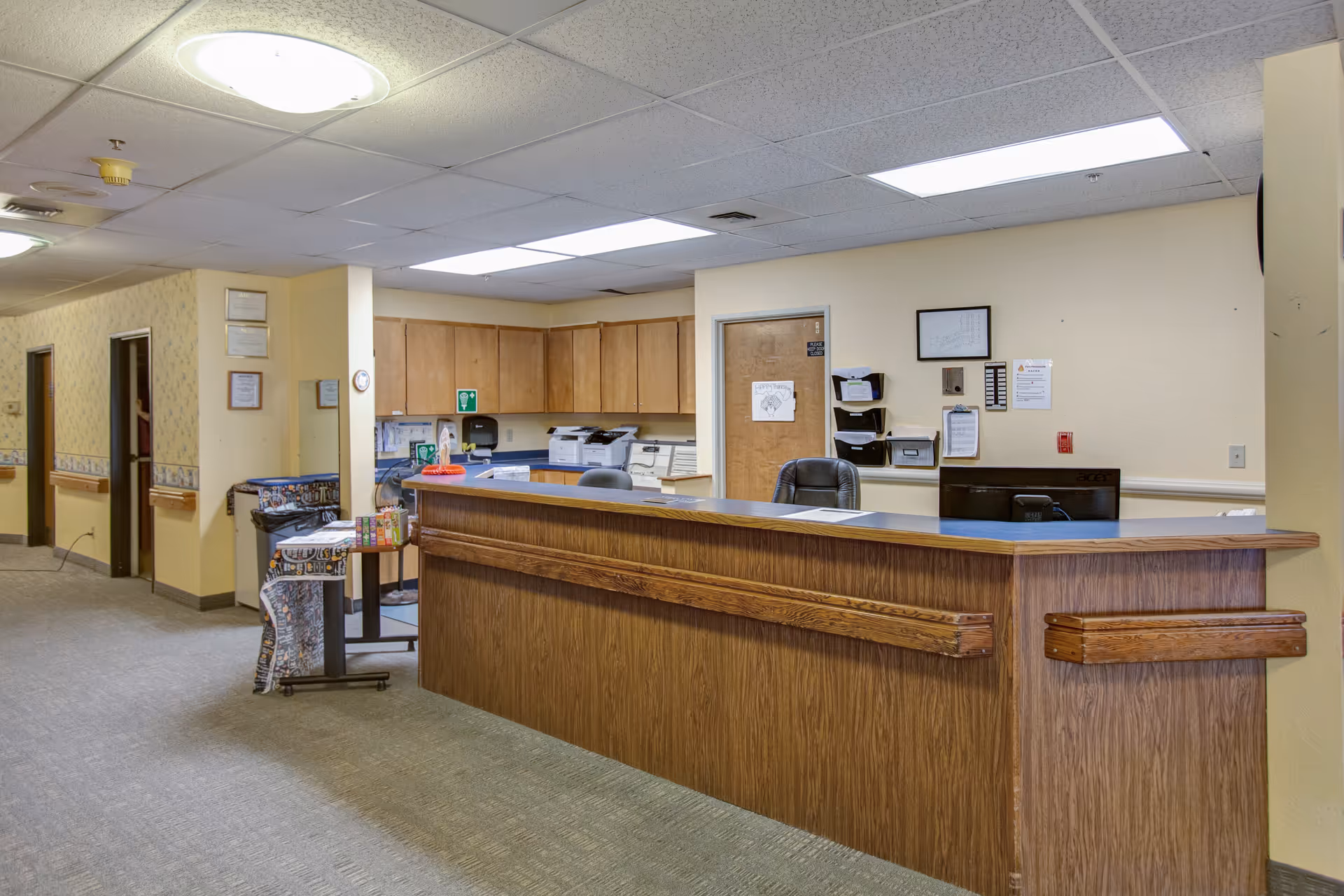 Reception desk area inside a senior living facility with wooden counter, office chairs, cabinets, and a hallway with doors leading to other rooms.