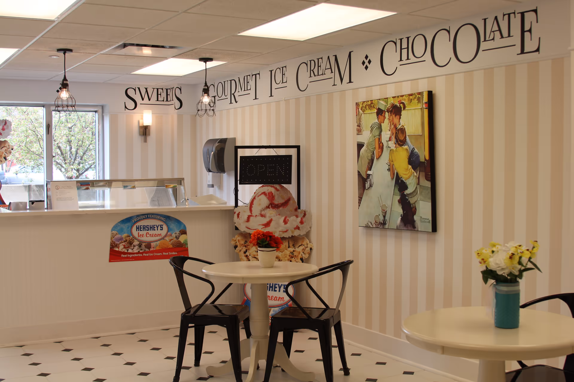 Interior view of a small ice cream parlor with two round tables, each with a flower vase on top. The walls have beige and white vertical stripes, and there is a large sign on the wall that reads 'Sweets Gourmet Ice Cream Chocolate.' There is a counter with a Hershey's Ice Cream sign and an open sign near the back. Two black metal chairs are placed around one of the tables.