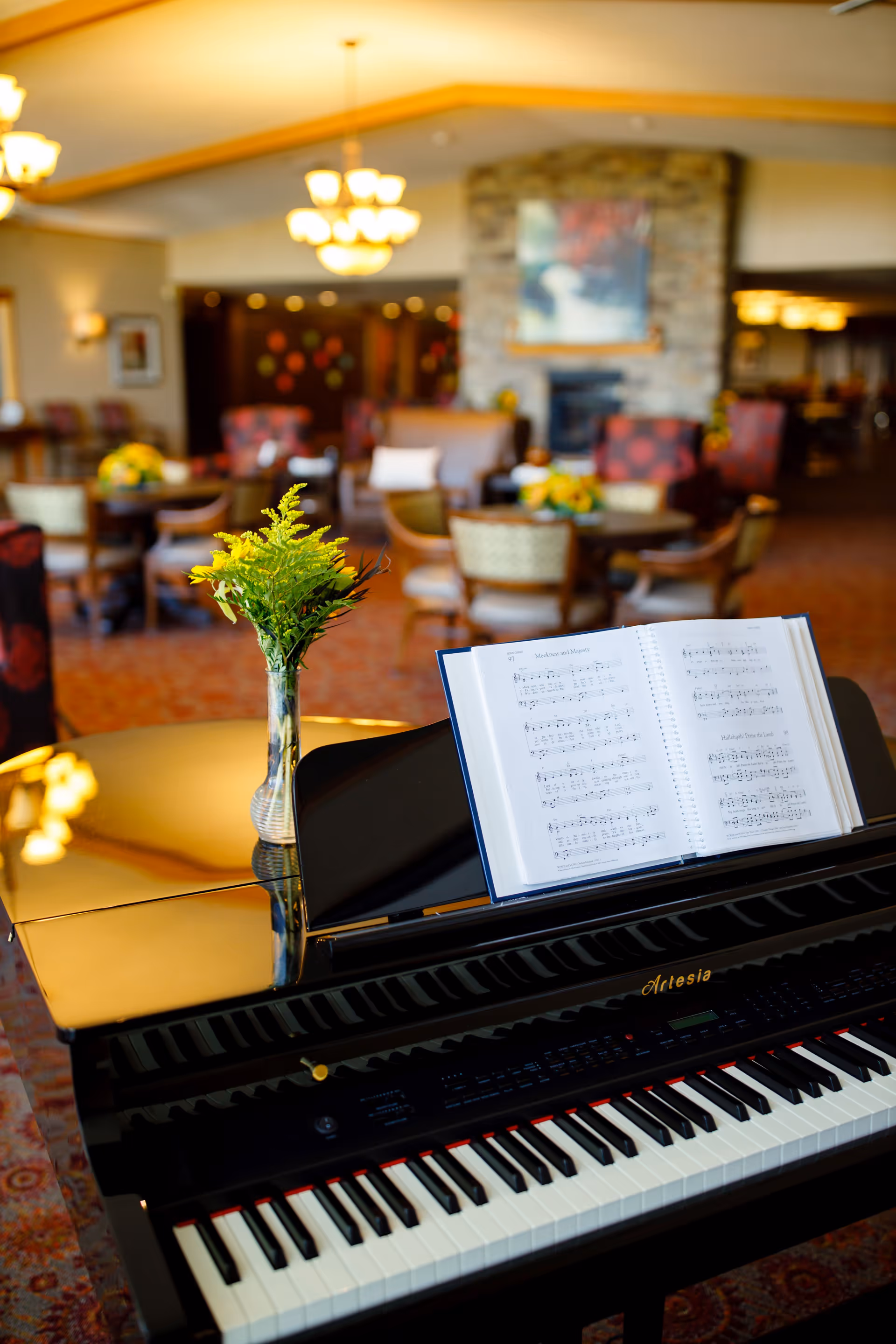 A black grand piano with an open music book and a small vase with green foliage on top, set in a warmly lit room with multiple round tables and chairs, a stone fireplace, and chandeliers hanging from the ceiling.