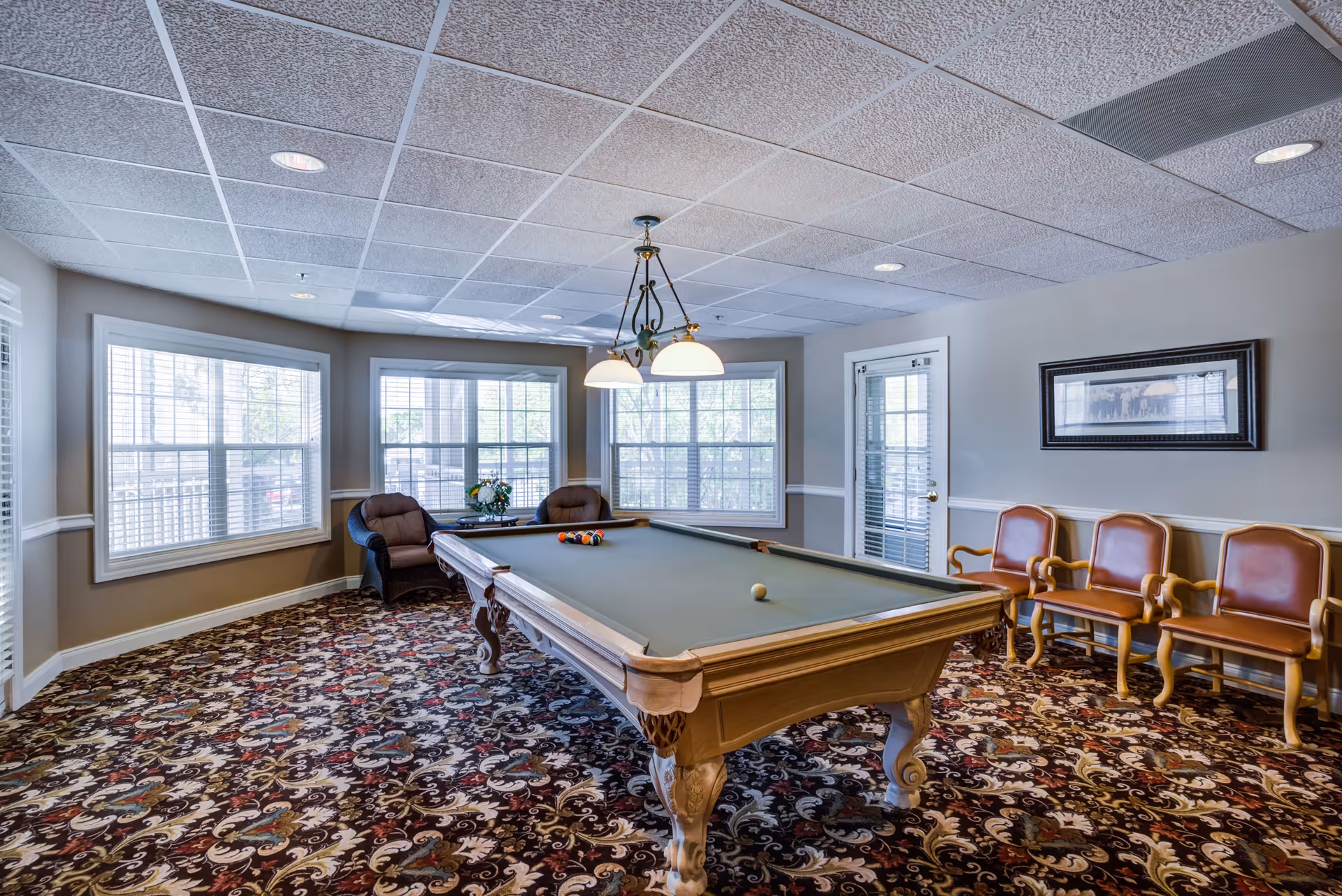 Bright recreation room with a pool table, seating chairs, patterned carpet and large windows.