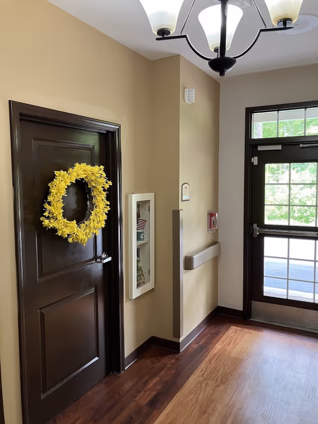 Interior hallway of Franklin Manor Assisted Living Center showing a dark wooden door decorated with a yellow floral wreath, a white emergency supply cabinet mounted on the beige wall, a fire alarm, a handrail, and a glass door leading outside with a view of greenery.