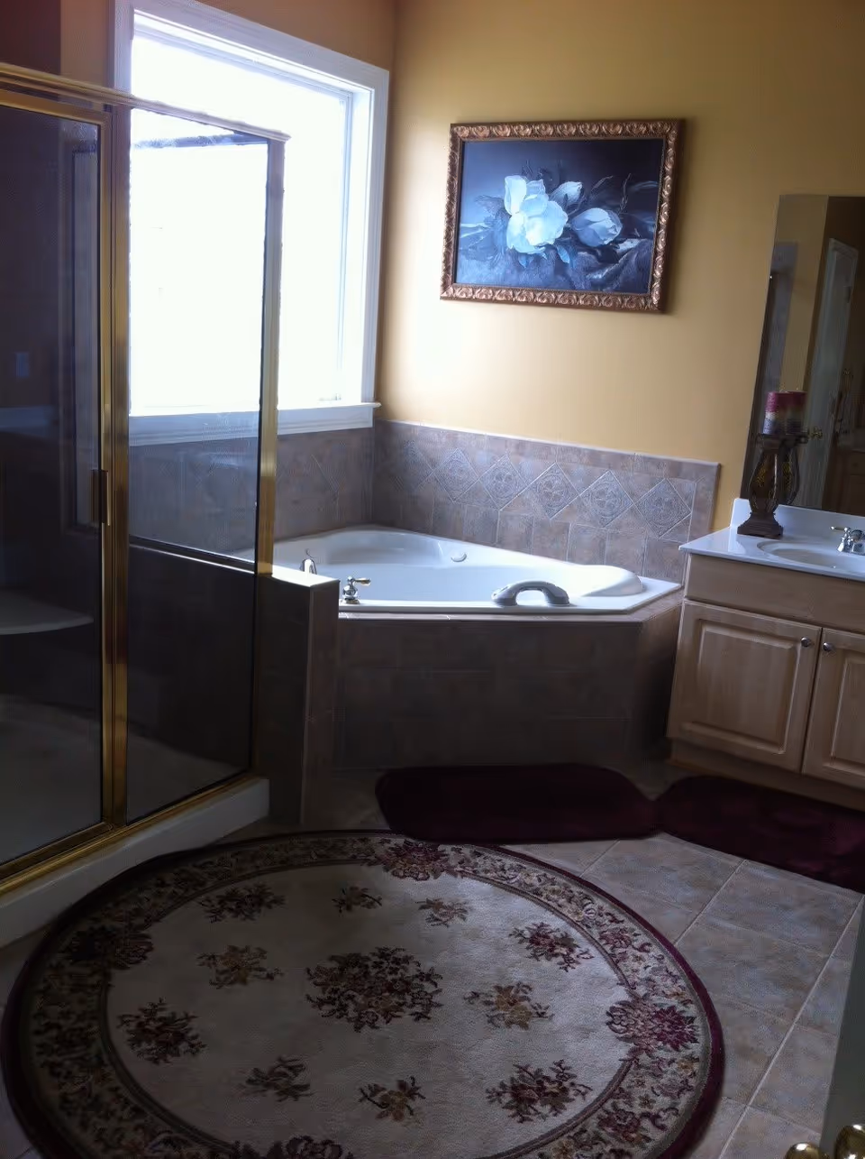 Bathroom with a corner bathtub surrounded by beige tiles, a glass-enclosed shower with gold trim, a vanity with a sink and cabinet, a large window letting in natural light, a framed floral painting on the yellow wall, and a round floral-patterned rug on the tiled floor.