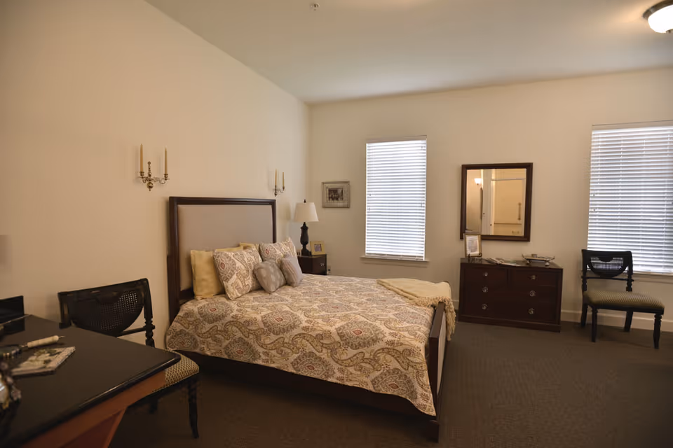 A neatly arranged bedroom with a double bed covered in a patterned quilt and multiple pillows. The room has two windows with blinds, a wooden dresser with a mirror above it, two chairs, a bedside table with a lamp, and wall-mounted candle holders.