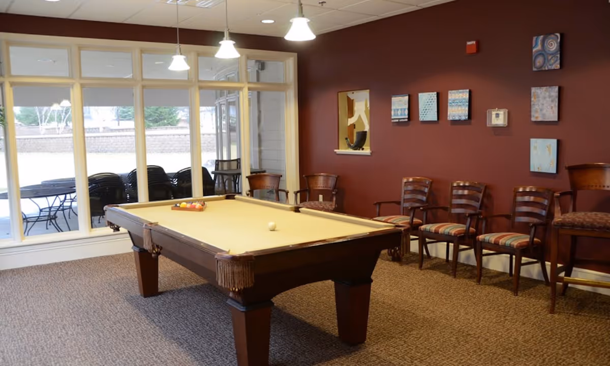 Game room interior with a pool table centered, chairs along a maroon wall, and large windows looking onto a patio.