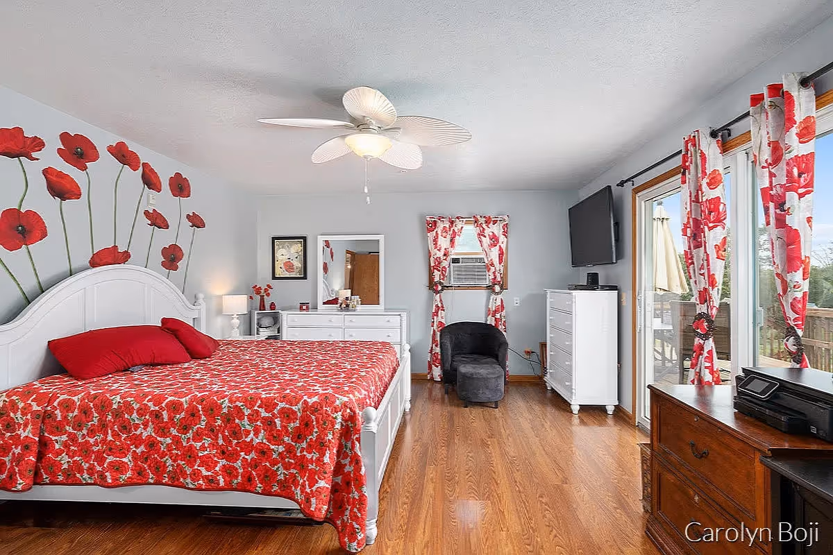 A bright bedroom with a white bed frame and a red floral bedspread. The wall behind the bed features a decorative mural of red poppies. There is a white dresser with a mirror, a black armchair, a white chest of drawers with a TV mounted above it, and a sliding glass door with red floral curtains leading to an outdoor area. The floor is wooden, and the ceiling has a white fan with a light.