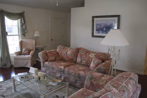 A living room with floral patterned sofa and armchair, a glass coffee table with candles and magazines, a floor lamp, a framed picture on the wall, and a window with green curtains.